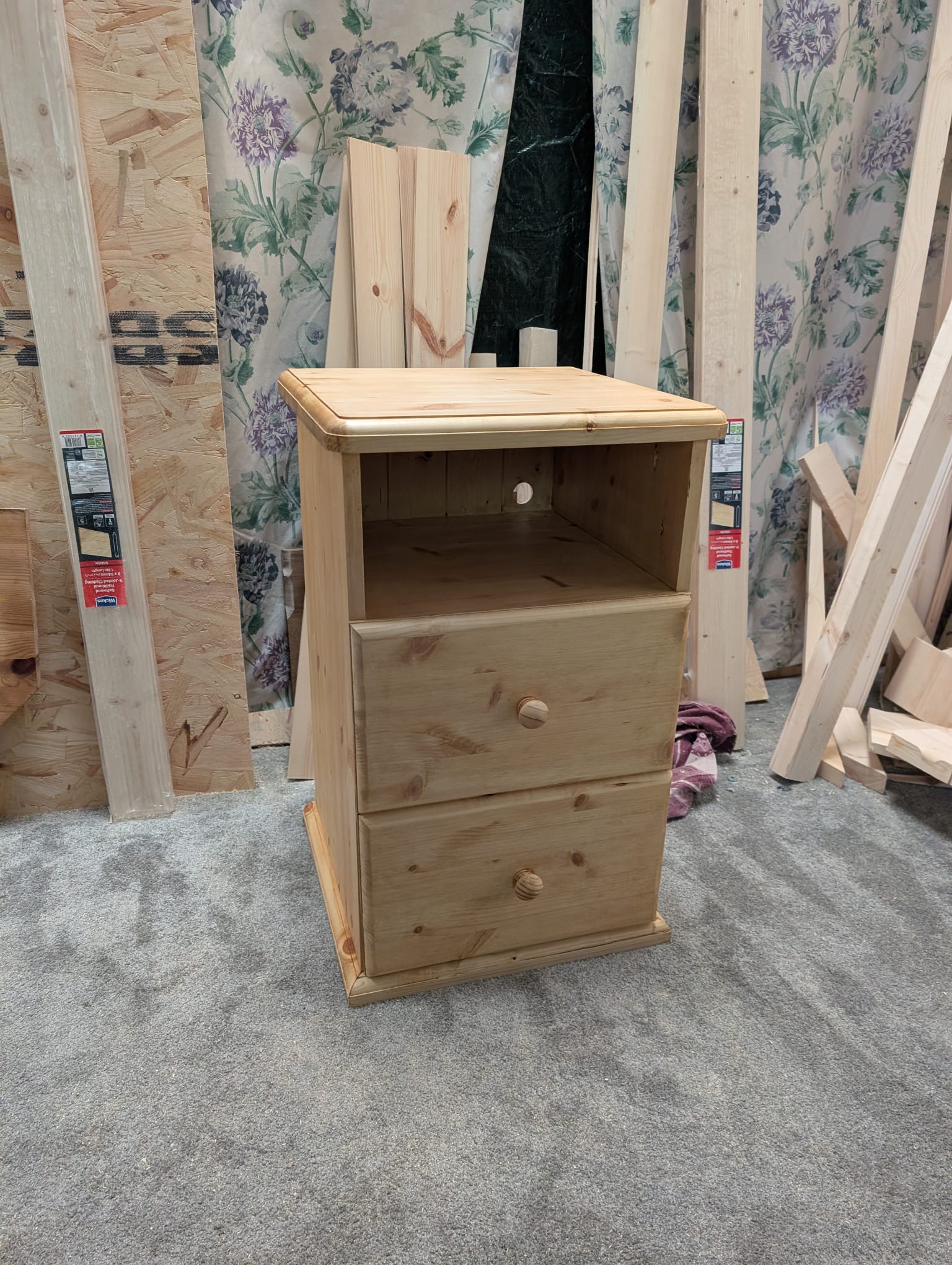 A single bedside cabinet stood in the workshop with both drawers closed, before oil, showing the chunky top, panelled sides and turned pine knobs.